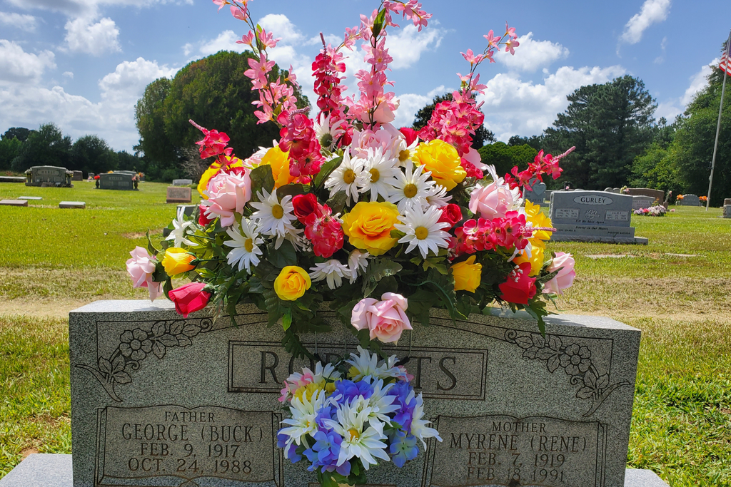 Colorful Floral Arrangement on Headstone
Silk headstone saddle with yellow roses, pink roses, daisies, and red tulips accented by lush greenery, displayed outdoors under a sunny sky.