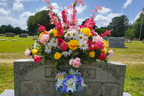 Colorful Floral Arrangement on Headstone
Silk headstone saddle with yellow roses, pink roses, daisies, and red tulips accented by lush greenery, displayed outdoors under a sunny sky.