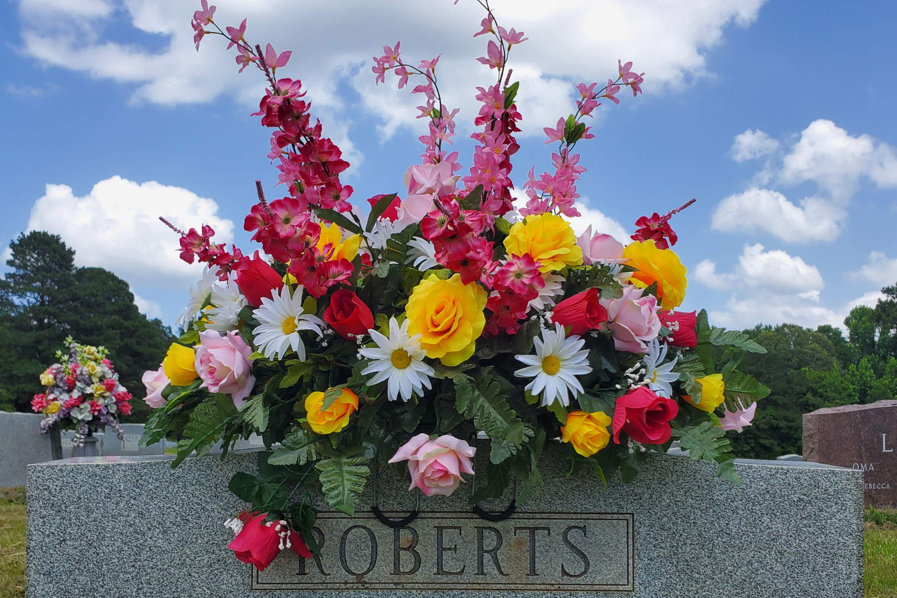 Bright Headstone Saddle with Mixed Florals
Artificial spring and summer headstone saddle featuring yellow roses, pink roses, white daisies, and red tulips with greenery on a granite memorial.