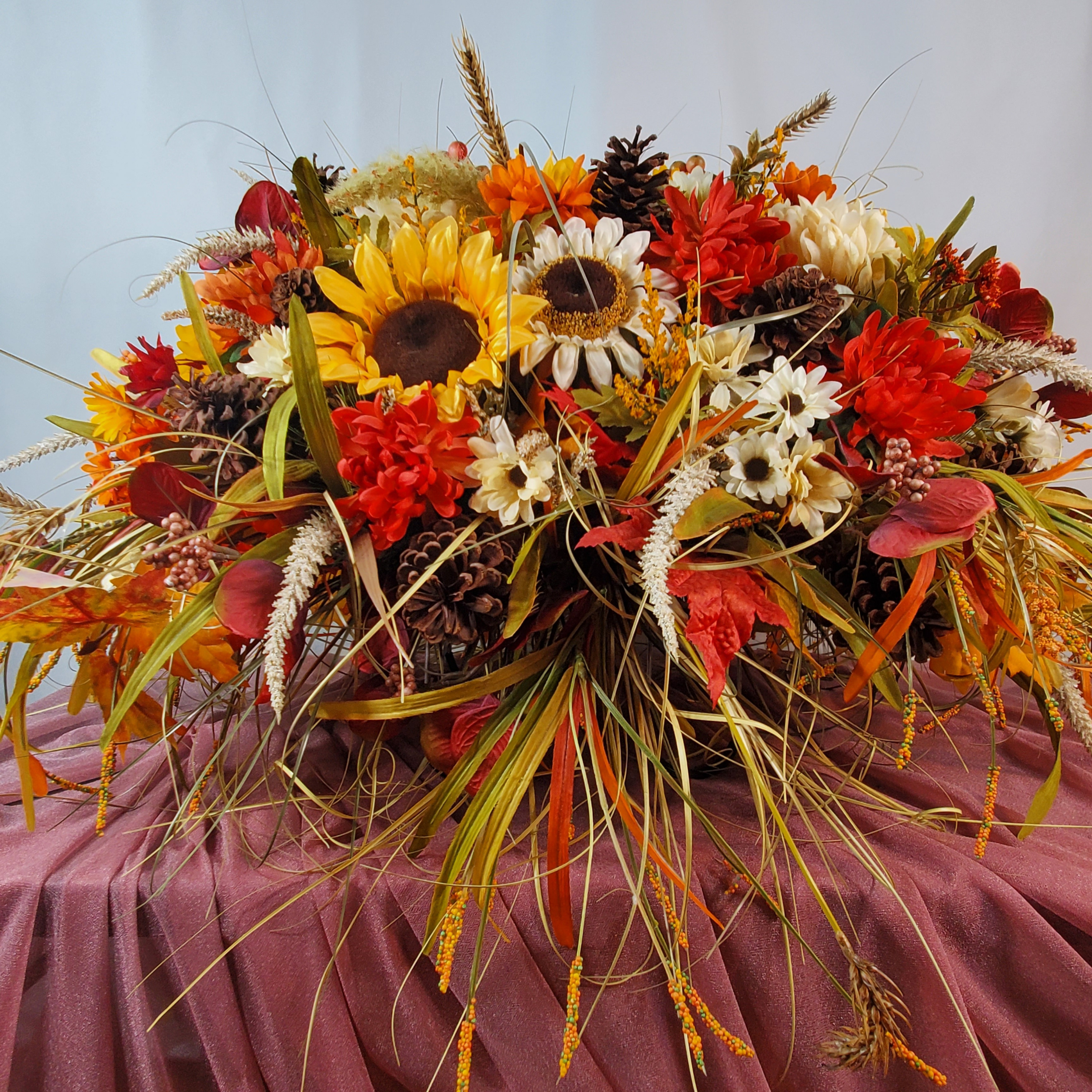Large artificial fall casket spray with sunflowers, maroon mums, golden leaves, and mixed greenery displayed atop a white casket outdoors.