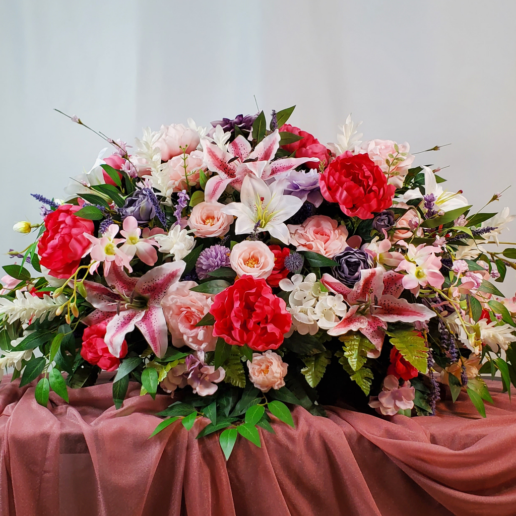 Spring Floral Headstone Saddle Arrangement
Artificial spring headstone saddle with pink lilies, red peonies, blush roses, lavender blooms, white hydrangeas, and lush greenery on a mauve draped background.